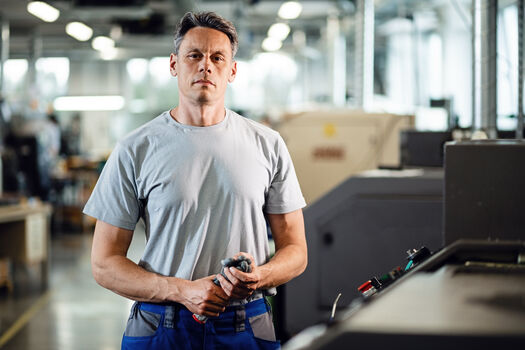 Ein Mann schaut in die Kamera, er hält Arbeitshandschuhe in der Hand. Im Hintergrund ist eine Werkshalle zu sehen mit vielen Maschinen.