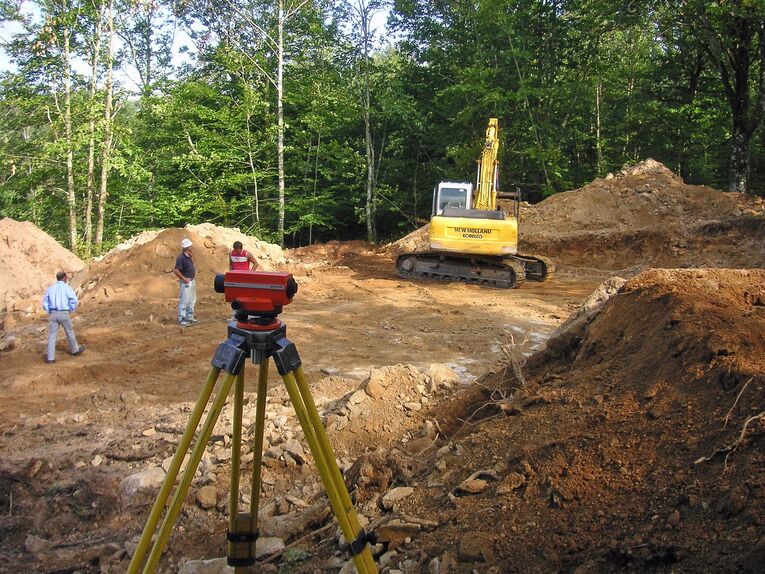 Eine Baustelle mit einem Bagger, im Vordergrund ein Vermessungsgerät im Hintergrund Menschen.