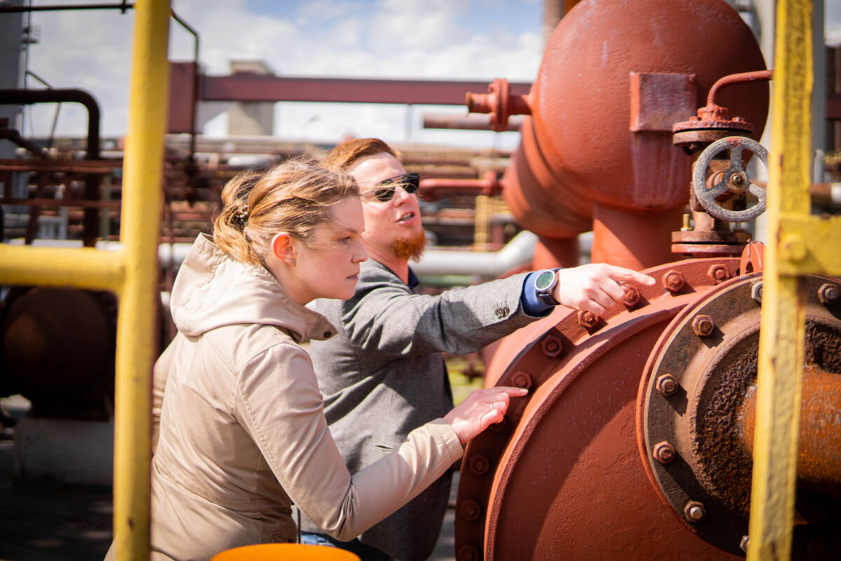 Zu sehen sind rostige Rohre und Rohrverbindungen und zwei Studierende, dei sich diese Bauteile prüfend anschauen. Beide zeigen auf eine Stelle am Rohr und sprechen miteinander. 
