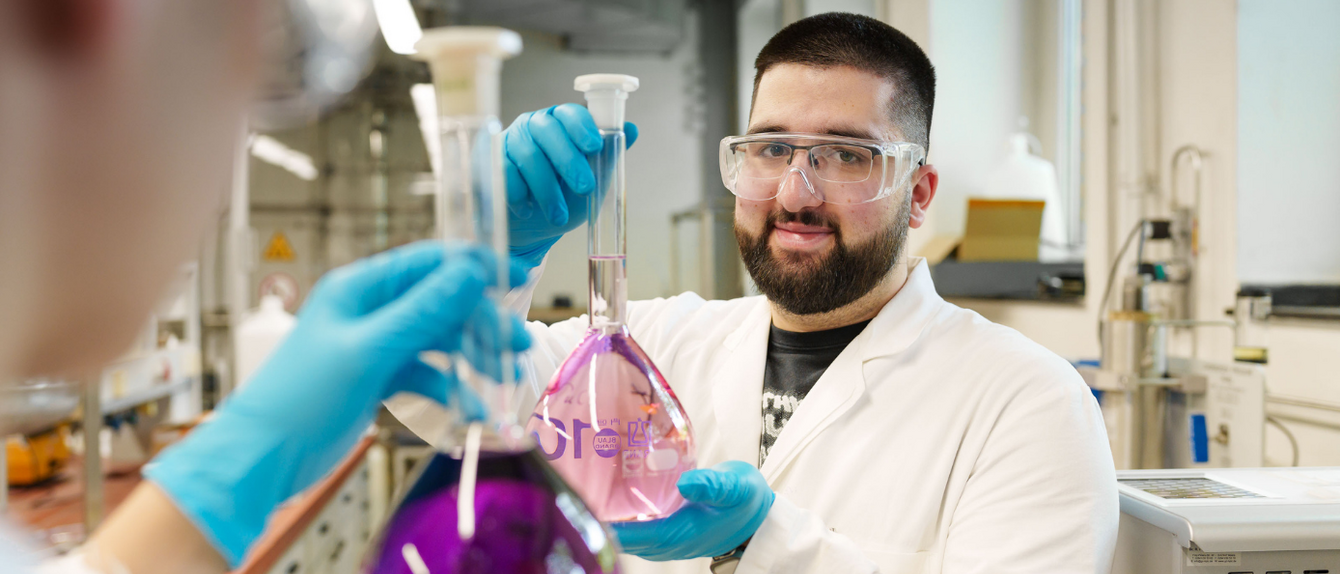 Ein Laborversuch im Labor für chemische Verfahrenstechnik. Glaskolben mit lila Flüssigkeit, dahinter ein Studierender, der in die Kamera blickt. 