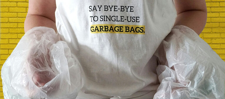 Ein junger Mensch trägt ein T-Shirt mit der englischen Aufschrift "Say bye-bye to single-use Garbage Bags" und hält zwei Plastiktüten in den Händen