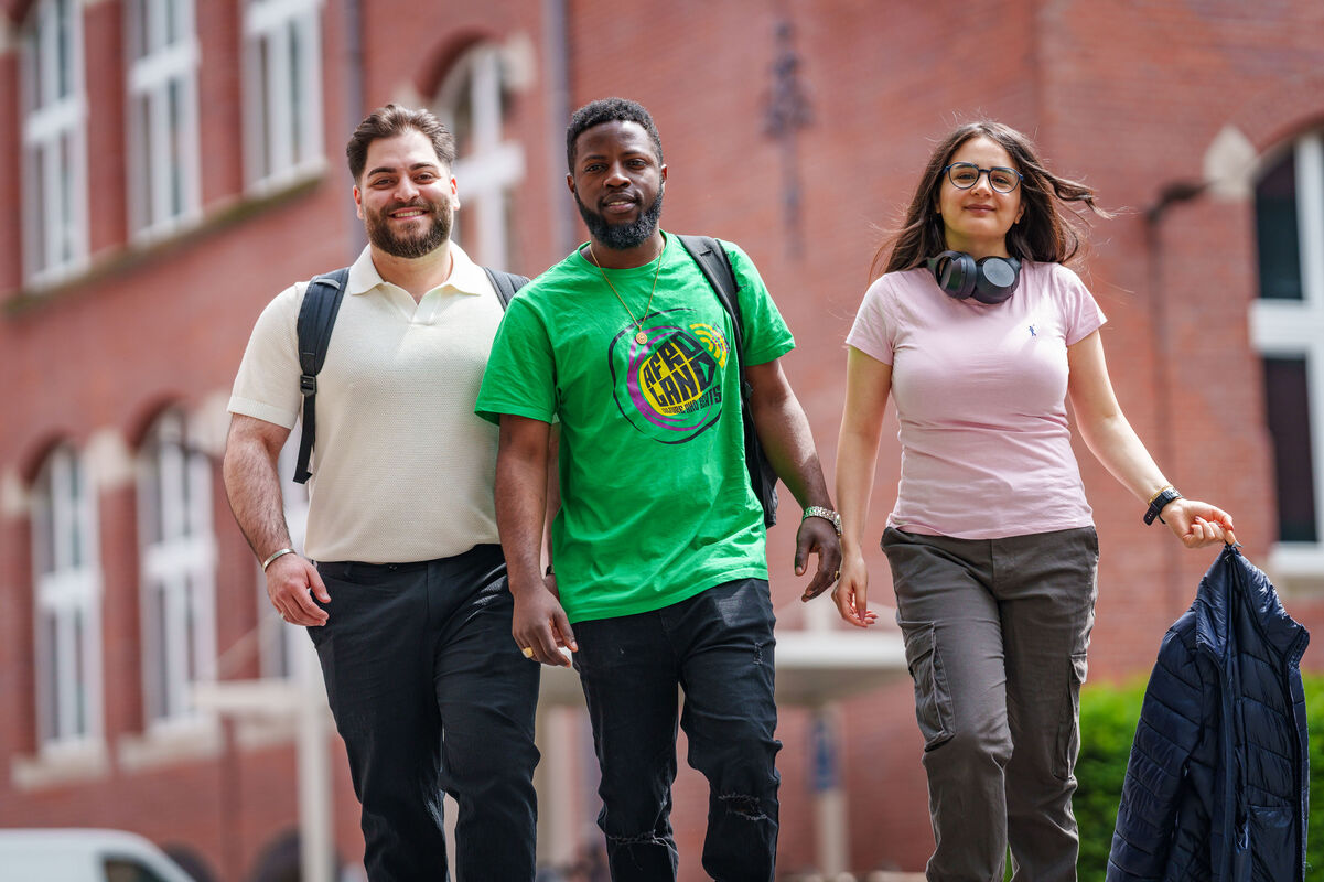 Zwei junge Männer und eine Frau laufen auf die Kamera zu, sie laufen über den Campus der THGA. 