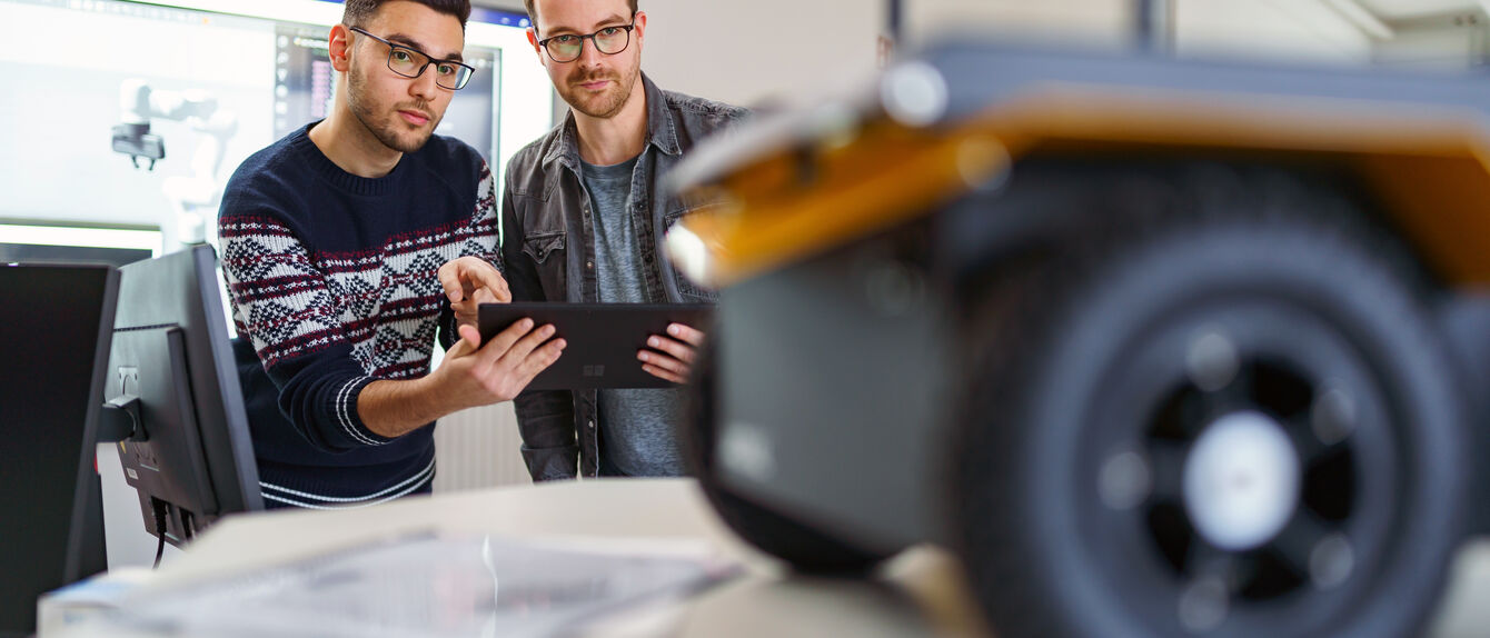 Ein Studierender und ein Mitarbeiter aus dem Fachbereich Elektrotechnik/Ingenieurinformatik sind im Robotiklabor der Hochschule und steuern einen fahrenden Roboter von Ferne. 