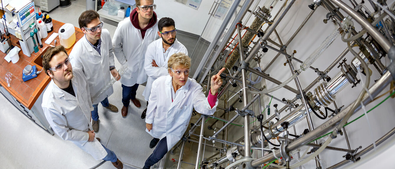 Vier Personen schauen sich ein sehr hohes technisches Laborgerät an.