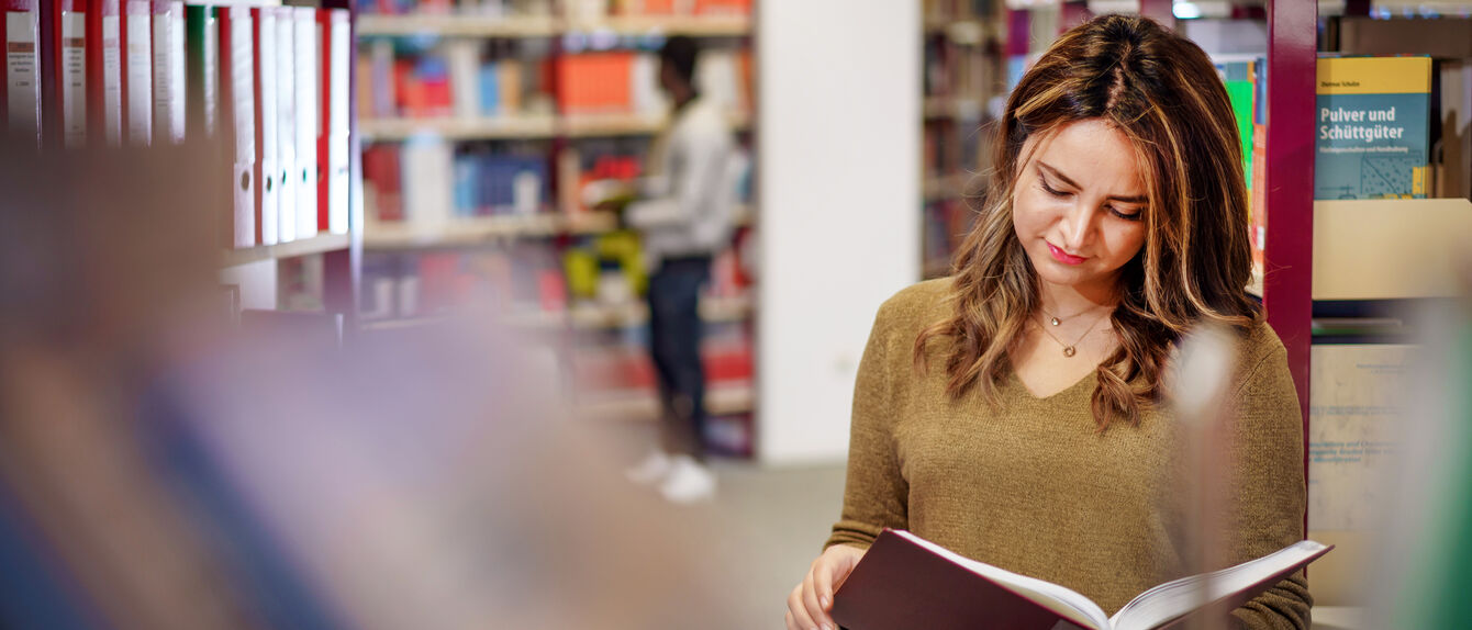 Eine Studentin steht in der Bibliothek der THGA und liest ein Buch über Ingenieurwesen.