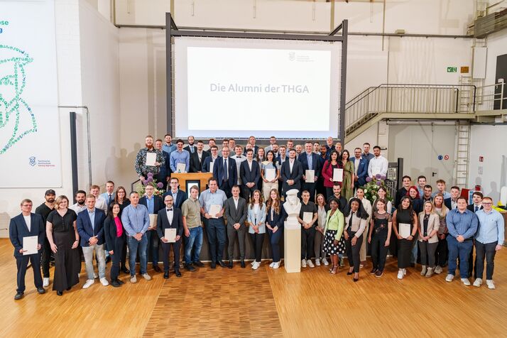 Alle Graduierten des Jahrgangs 2025 stehen auf und um die Bühne zum Gruppenfoto im Studierendenzentrum der THGA versammelt. 