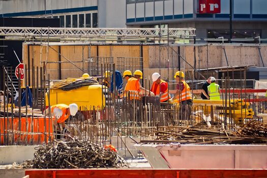 Auf dem Bild ist eine Großbaustelle zu sehen, im Hintergrund ist eine Gruppe Bauarbeiter mit eine Projektleiter zu sehen. Alle tragen Warnwesten und Helme und schauen interessiert in die Mitte auf einen Plan.