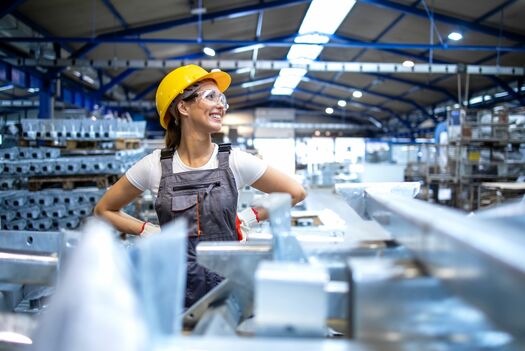 Eine Frau, die in einer Produktionshalle steht und zur Seite blickt. Sie ist mit Schutzhelm und -brille ausgestattet.