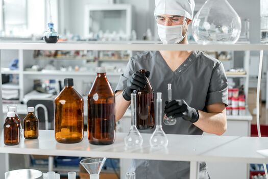 Eine Person steht hinter einem Regal mit Laborflaschen und prüft etwas. 