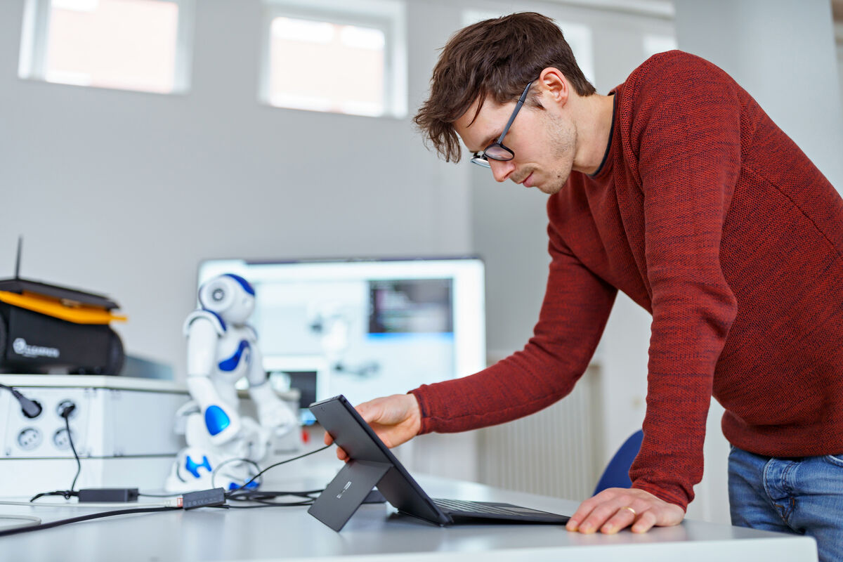Ein Informatik-Student blickt und tippt auf seinem Laptop im Robotiklabor. Im Hintergrund ist ein Roboter zu sehen. 