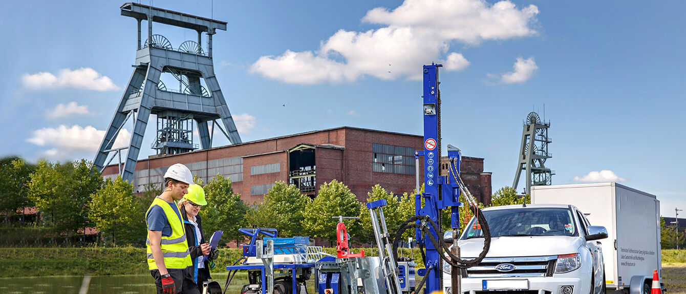 Zu sehen ist ein ehemaliges Zechengelände mit zwei Fördertürmen. Eine Studierende und ein Wissenschaftler schauen auf ein Clipboard. Rechts von beiden ist ein Bohrgerät zu sehen, daneben steht ein Auto der Technischen Hochschule Georg Agricola. 
