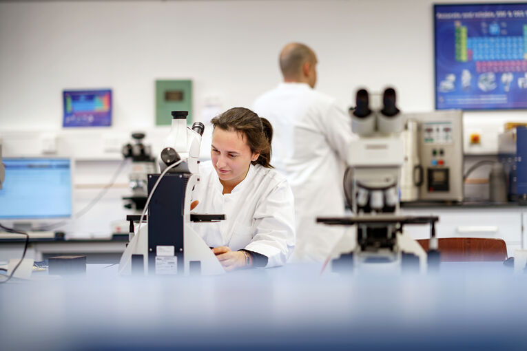Eine Professorin untersucht mit dem Mikroskop Materialien. Im Hintergrund steht ein Wissenschaftler, der einen materialwissenschaftliche Auswertung betrachtet.