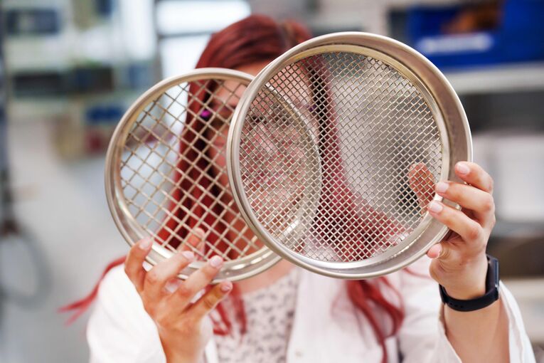 Im Labor der mechanischen Verfahrenstechnik hält eine Frau zwei Siebe für unterschiedliche Korngrößen vor ihr Gesicht.