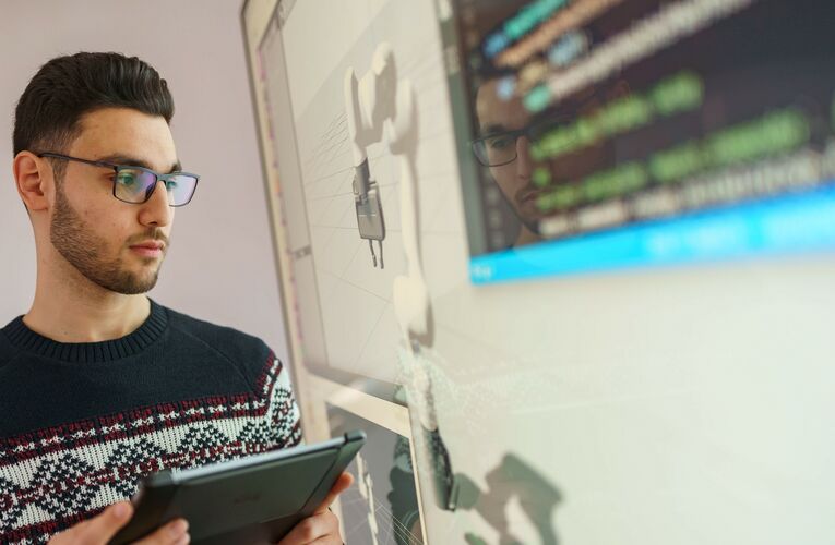 Ein Student steht vor einem Whiteboard. Er schaut darauf und man sieht dort ein Roboterarm und technische Zeichnungen. Der Student hält ein Tablet in der Hand. 