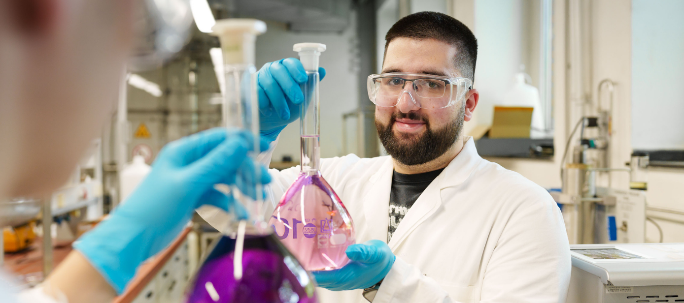 Ein Laborversuch im Labor für chemische Verfahrenstechnik. Glaskolben mit lila Flüssigkeit, dahinter ein Studierender, der in die Kamera blickt. 