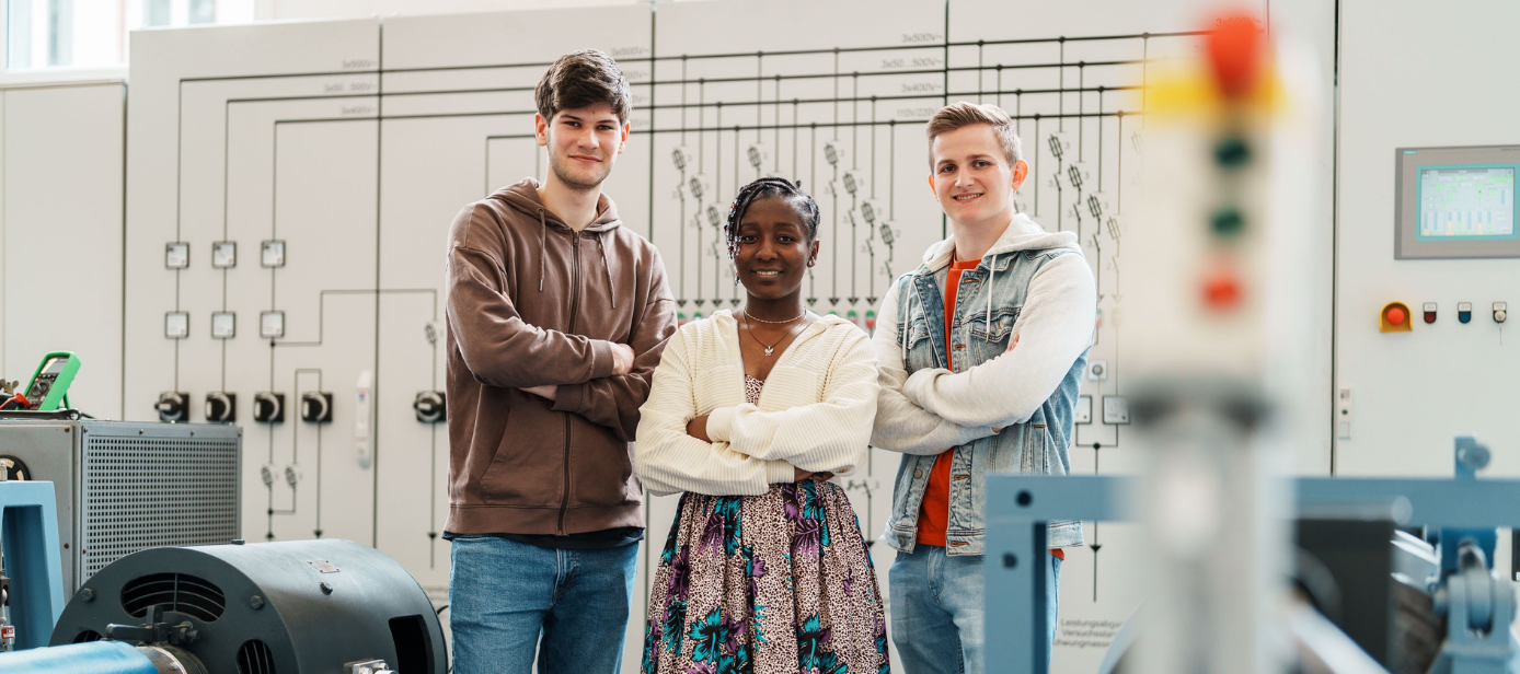 Drei Studierende der Elektrotechnik schauen zufrieden in die Kamera. In der Mitte steht eine weibliche Studierende. Labor für Leistungselektronik an der THGA. 