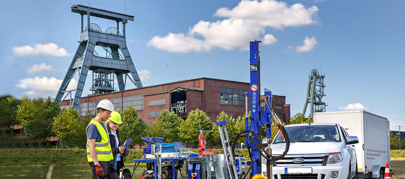 Zu sehen ist ein ehemaliges Zechengelände mit zwei Fördertürmen. Eine Studierende und ein Wissenschaftler schauen auf ein Clipboard. Rechts von beiden ist ein Bohrgerät zu sehen, daneben steht ein Auto der Technischen Hochschule Georg Agricola. 